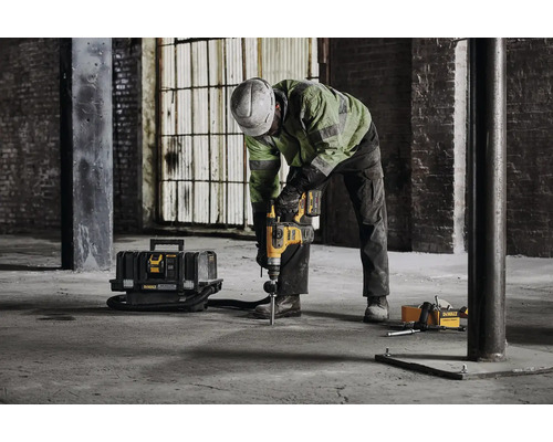 Stavební dělník vrtá vrtacím kladivem díru do betonové podlahy v průmyslové budově. Poblíž je box na nářadí a vysavač.