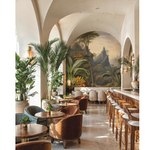 Světlý bar s klenutými stropy, tropickou nástěnnou malbou, dřevěným nábytkem, sametovými křesly a velkými palmami v květináčích.