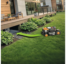 Robotická sekačka na trávu jede po zelené ploše ke své nabíjecí stanici pod dřevěnou terasou.