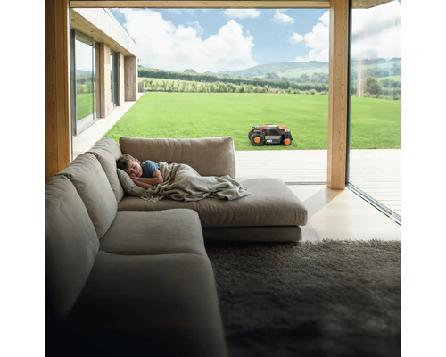 Chlapec spí na gauči, zatímco robotická sekačka pracuje na zahradě