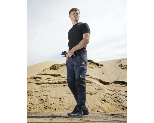 Muž v pracovním oděvu s pracovními kalhotami, tričkem a bezpečnostní obuví před písečným kopcem