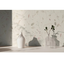 Světlá tapeta s jemným květinovým vzorem a trávami, před kterou stojí bílá keramická váza a dvě skleněné vázy na světlém povrchu.