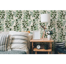 Vliesová tapeta se vzorem zelených listů ve světlé ložnici s dřevěným stolem, lampou a polštáři.