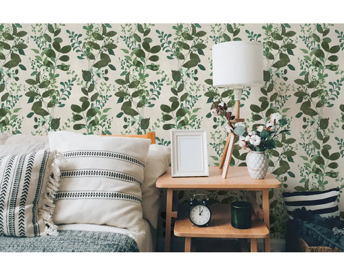 Vliesová tapeta se vzorem zelených listů ve světlé ložnici s dřevěným stolem, lampou a polštáři.