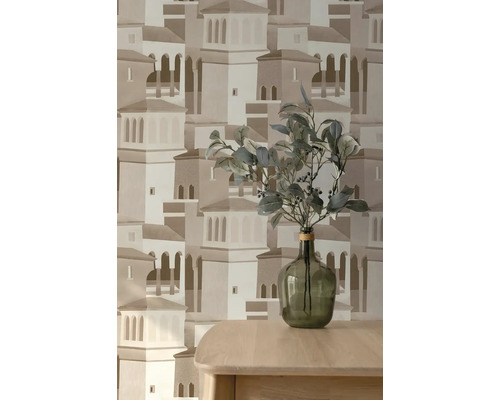 Vliesová tapeta s architektonickým vzorem středomořského města, před ní dřevěný stůl se skleněnou vázou a zelenými větvemi.