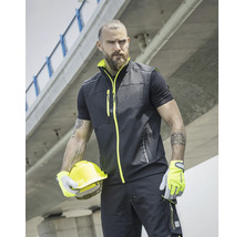 Muž má na sobě pracovní vestu, tričko, pracovní kalhoty, rukavice a drží ochrannou přilbu.