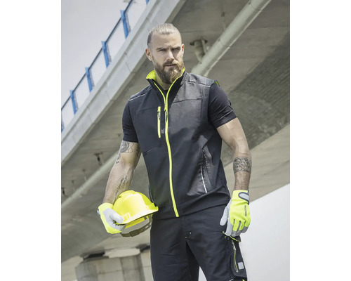 Muž má na sobě pracovní vestu, tričko, pracovní kalhoty, rukavice a drží ochrannou přilbu.