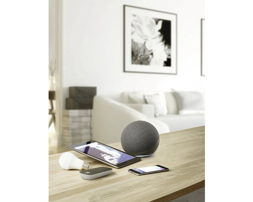 Snímek obývacího pokoje s chytrými domácími zařízeními, jako jsou reproduktory, tablety, smartphony a žárovky na stole.