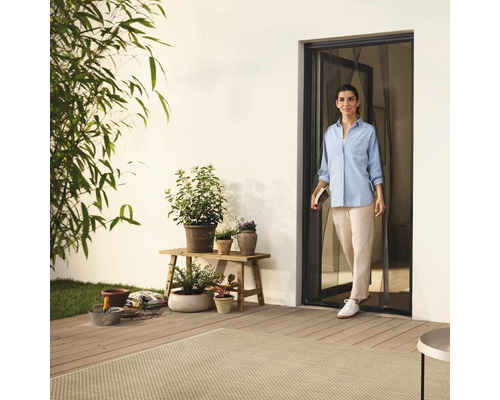 Žena vstupuje na terasu dveřmi se sítí proti hmyzu, obklopena rostlinami a zahradním příslušenstvím.