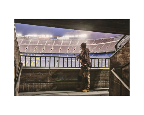 Muž v pracovním oděvu stojí před stadionem a dívá se na hřiště.