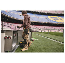 Muž s opaskem na nářadí a pracovním oděvem pracující na elektrické skříni na stadionu.