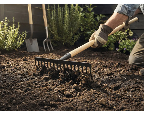 Zahradník hrábě zeminu hráběmi, v pozadí je vidět rýč a vidle.