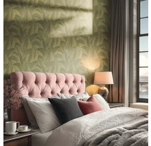 Útulná ložnice s tapetou se zeleným vzorem listů, růžovým čalouněným čelem postele a hřejivým osvětlením ze stolní lampy.