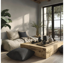 Světlý obývací pokoj s masivním dřevěným stolem, nízkou pohovkou, polštáři a velkými okny v minimalistickém stylu s přírodní úpravou stěn.