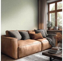 Obývací pokoj se strukturovanou světle zelenou tapetou, hnědou koženou pohovkou s polštáři, dřevěným oknem a světlou, útulnou atmosférou.