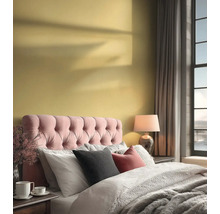 Světlá ložnice se žlutou stěnou, růžovým polstrovaným čelem postele, šedým povlečením a lampičkou u velkého okna.