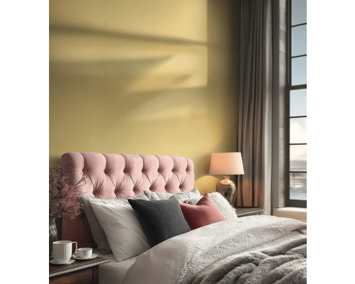 Světlá ložnice se žlutou stěnou, růžovým polstrovaným čelem postele, šedým povlečením a lampičkou u velkého okna.