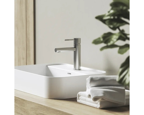 Moderní umyvadlová baterie ve vzhledu nerezové oceli na bílém umyvadle na desku, na dřevěné desce vedle ručníků ve světlé koupelně.