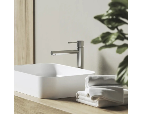 Moderní koupelna s vysokou pákovou baterií z kartáčovaného kovu a bílým obdélníkovým umyvadlem na dřevěné desce s ručníky.