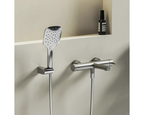 Moderní chromová sprchová souprava skládající se z hranaté ruční sprchy v nástěnném držáku a válcové sprchové baterie se sprchovou hadicí.