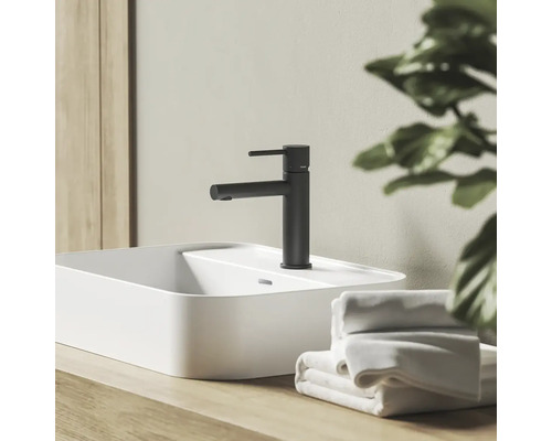 Černá matná umyvadlová baterie na bílém umyvadle na desku na dřevěném podkladu v koupelně s ručníky.