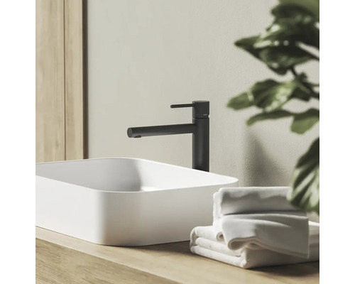 Moderní koupelnová scéna s černou pákovou umyvadlovou baterií a bílým umyvadlem na desku na dřevěném podkladu s ručníky.