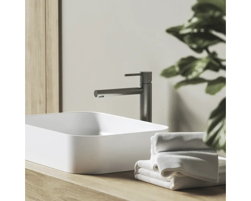 Moderní koupelna s bílým umyvadlem na desku, tmavě šedou baterií na dřevěné desce a bílými ručníky.