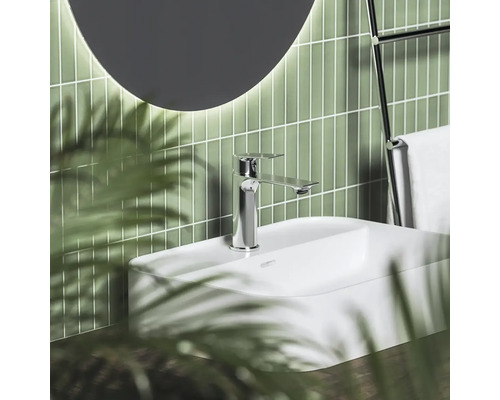 Moderní koupelna s bílým umyvadlem, chromovou baterií, zelenými vertikálními obklady a kulatým zrcadlem.