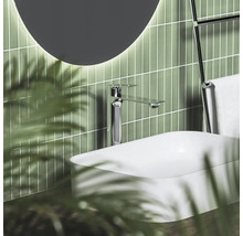 Moderní umyvadlová baterie v chromu na bílém umyvadle na desku před zelenými svislými obklady s kulatým osvětleným zrcadlem.