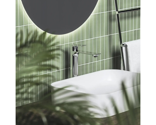 Moderní umyvadlová baterie v chromu na bílém umyvadle na desku před zelenými svislými obklady s kulatým osvětleným zrcadlem.