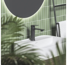 Černá matná umyvadlová baterie na bílém umyvadle před zelenou obloženou stěnou s kulatým zrcadlem a držákem na ručníky v moderní koupelně.