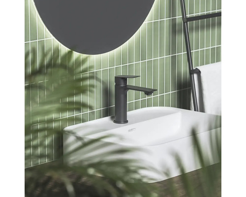 Černá matná umyvadlová baterie na bílém umyvadle před zelenou obloženou stěnou s kulatým zrcadlem a držákem na ručníky v moderní koupelně.