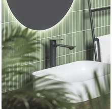 Černá umyvadlová baterie u bílého umyvadla na desku před zelenými obklady s osvětleným zrcadlem v moderní koupelně.