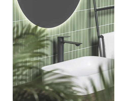 Černá umyvadlová baterie u bílého umyvadla na desku před zelenými obklady s osvětleným zrcadlem v moderní koupelně.