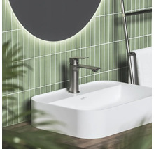 Moderní koupelna s bílým umyvadlem na desku, kovovou baterií, zelenými svislými obklady a kulatým podsvíceným zrcadlem.