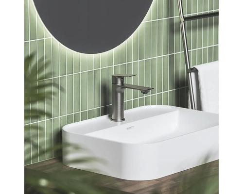 Moderní koupelna s bílým umyvadlem na desku, kovovou baterií, zelenými svislými obklady a kulatým podsvíceným zrcadlem.