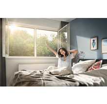 Světlá ložnice s dokořán otevřeným plastovým oknem, kterým proniká sluneční světlo, a osobou v posteli, která se při probuzení protahuje.