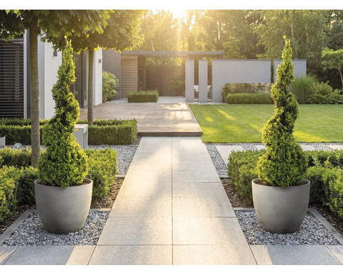 Moderní světlá zahrada s kamennou cestou, spirálovitě tvarovanými dřevinami v kulatých květináčích, dřevěnou terasou a trávníkem.