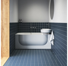 Moderní koupelna s bílou vanou, nad ní namontovaným umyvadlem a tmavě modrými čtvercovými obklady na stěně a podlaze.