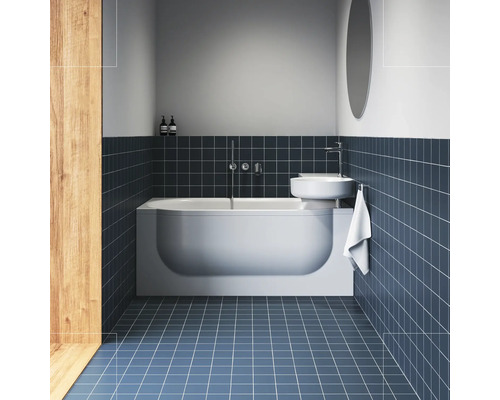 Moderní koupelna s bílou vanou, nad ní namontovaným umyvadlem a tmavě modrými čtvercovými obklady na stěně a podlaze.