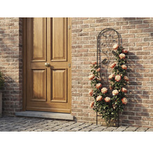 Kovová mříž na popínavé rostliny s motivem ptáků a listů, s pnoucími růžemi u cihlové stěny vedle dřevěných dveří.