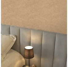 Strukturovaná béžová tapeta nad šedým čalouněným čelem postele v ložnici s rozsvícenou noční lampou.