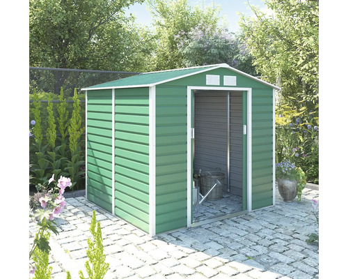 Zelený kovový zahradní domek se sedlovou střechou a bílým rámem, posuvnými dveřmi a ventilací, na kamenné dlažbě ve světlé zahradě.