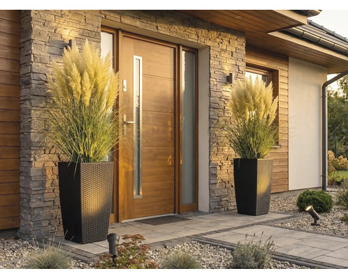 Moderní vchod do domu s dřevěnými dveřmi, kamenným obkladem a dvěma vysokými květináči v ratanovém vzhledu s pampovou trávou.