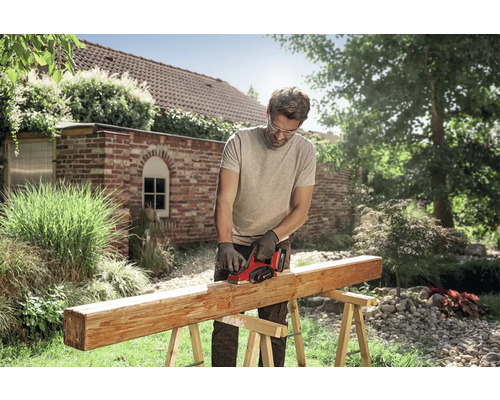 Muž opracovává dřevěný trám na kozách elektrickým hoblíkem v jasné zahradě. Má na sobě ochranné brýle a rukavice.