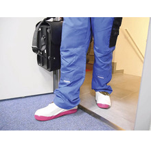 Osoba v modrých pracovních kalhotách s taškou na nářadí má na sobě bílé návleky na obuv s růžovou podrážkou pro ochranu podlahy.