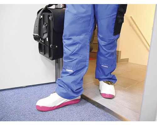 Osoba v modrých pracovních kalhotách s taškou na nářadí má na sobě bílé návleky na obuv s růžovou podrážkou pro ochranu podlahy.