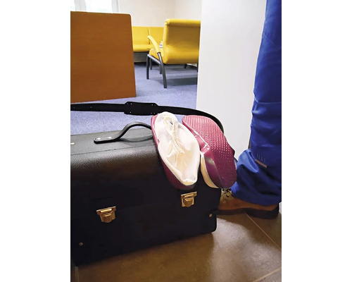 Černá brašna na nářadí s červenými návleky na obuv ve světlé místnosti se žlutými křesly a osobou v modrých pracovních kalhotách.