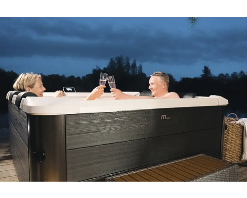 Pár relaxuje v obdélníkové vířivce MSpa s tmavým venkovním obložením v dekoru dřeva na zahradě za soumraku.