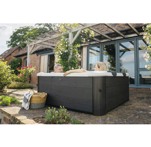 Dvě osoby relaxují ve čtvercové venkovní vířivce s tmavě šedým dřevěným obložením na terase u domu s pergolou.